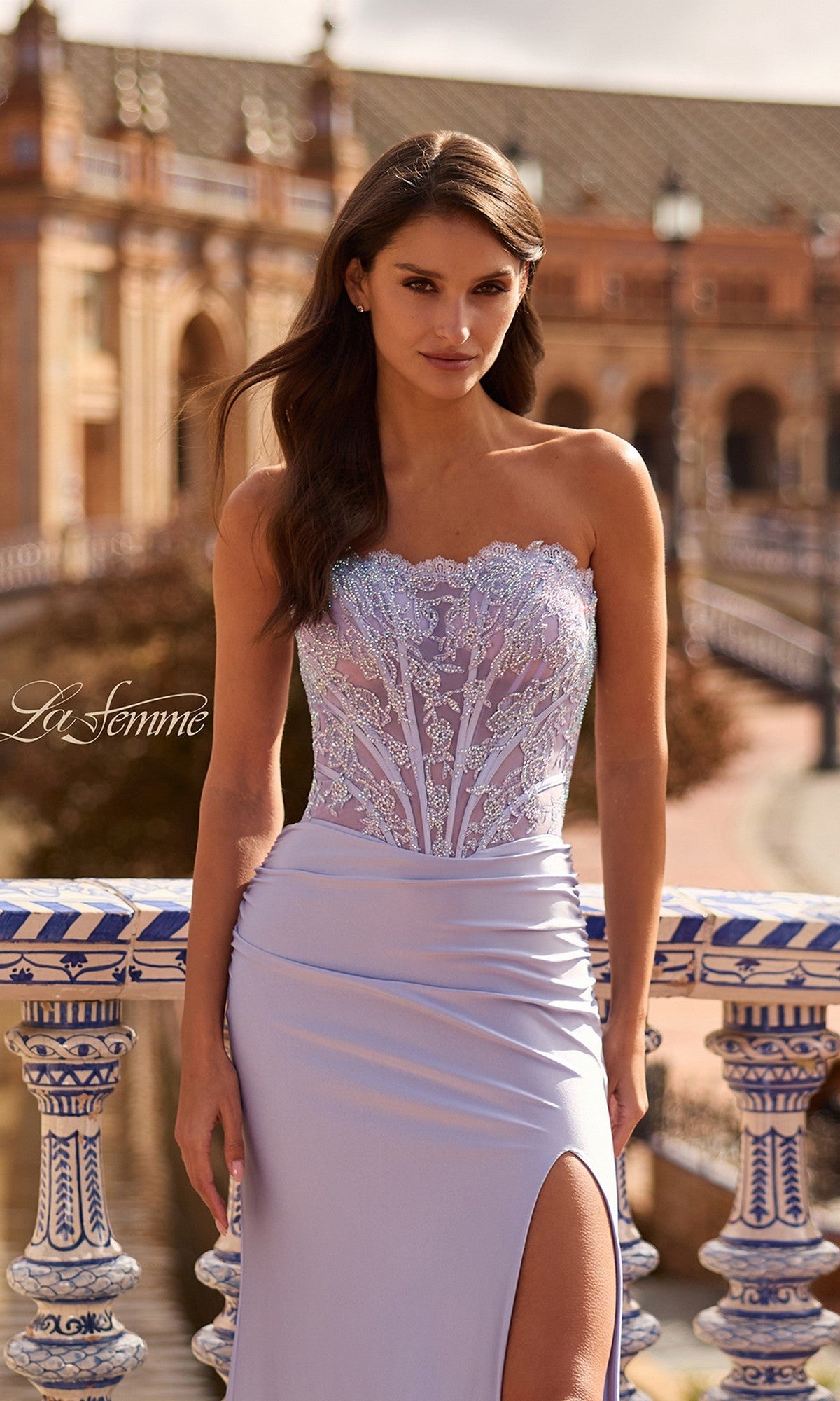 A woman stands outdoors by ornate railings in a La Femme 33509 long prom dress, featuring a strapless mermaid silhouette, beaded lace corset, and thigh-high slit, with historic architecture in the background.